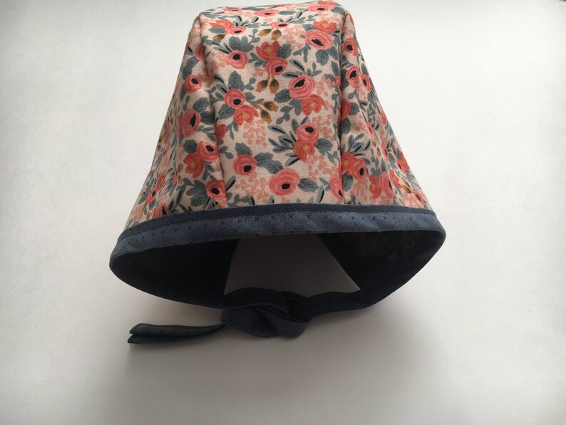 May include: A floral print surgical cap with a navy blue trim and tie. The cap features a pink and white floral pattern.