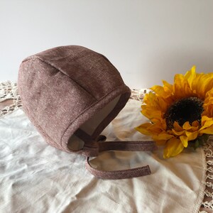 May include: A brown knitted baby bonnet with a tie. The bonnet is on a white fabric surface with a crocheted lace trim. A yellow sunflower is in the background.