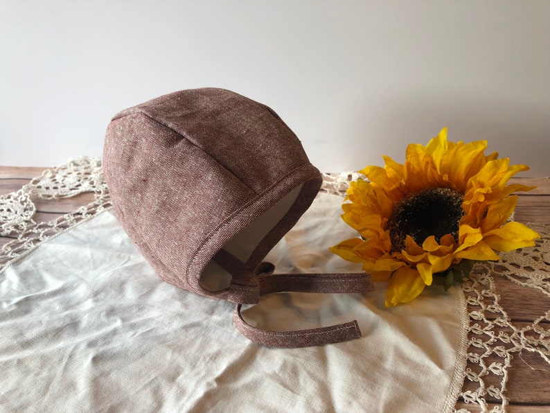 May include: A brown knitted baby bonnet with a tie. The bonnet is on a white fabric surface with a crocheted lace trim. A yellow sunflower is in the background.