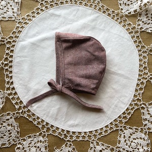 May include: A mauve bonnet with a tie, made of a soft, textured fabric. The bonnet is lying on a white doily with a crocheted lace border.
