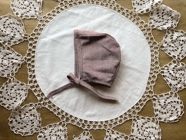 May include: A mauve bonnet with a tie, made of a soft, textured fabric. The bonnet is lying on a white doily with a crocheted lace border.