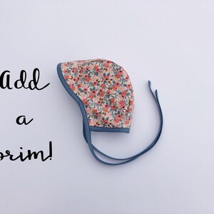 May include: A baby bonnet with a floral print and blue ties. The text "Add a brim!" is written in black on a white background.