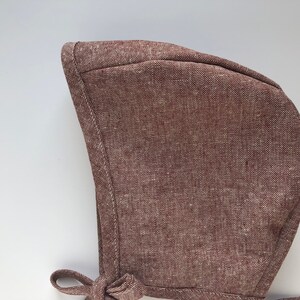 May include: A brown linen baby bonnet with a tie. The bonnet is made of a soft, textured fabric.