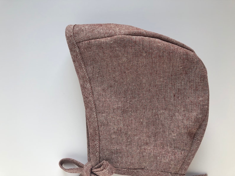 May include: A brown linen baby bonnet with a tie. The bonnet is made of a soft, textured fabric.