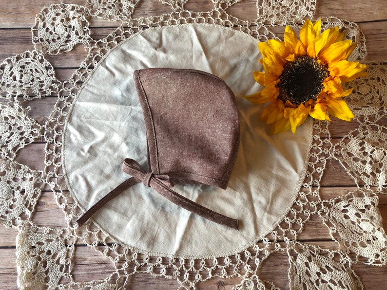 May include: A brown linen baby bonnet with a bow tie. The bonnet is on a white doily with a crocheted edge.