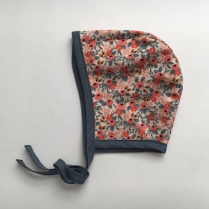 May include: A floral baby bonnet with a pink and red floral print and a navy blue trim. The bonnet has a tie at the back.
