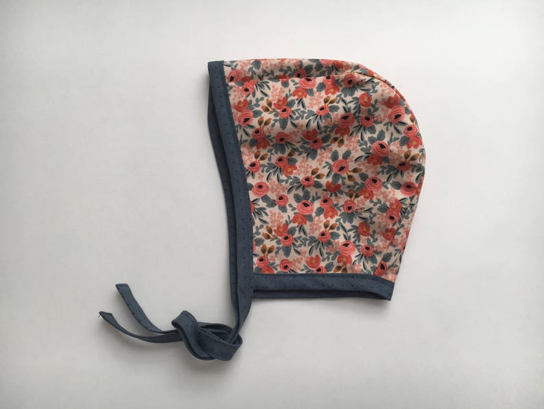 May include: A floral baby bonnet with a pink and red floral print and a navy blue trim. The bonnet has a tie at the back.