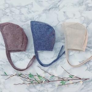 May include: Three baby bonnets in different colors: brown, blue, and beige. The bonnets are made of a soft, textured fabric and have long ties.