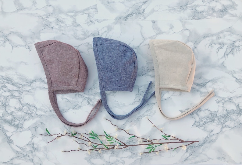 May include: Three baby bonnets in different colors: brown, blue, and beige. The bonnets are made of a soft, textured fabric and have long ties.