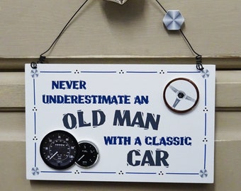 May include: White wooden sign with blue text that reads "Never underestimate an old man with a classic car". The sign has three round black and white gauges and a steering wheel.
