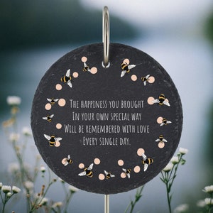 May include: A black slate garden stone with a white inscription that reads "THE HAPPINESS YOU BROUGHT IN YOUR OWN SPECIAL WAY WILL BE REMEMBERED WITH LOVE EVERY SINGLE DAY." The stone is decorated with a circle of pink dots and yellow and black bees.