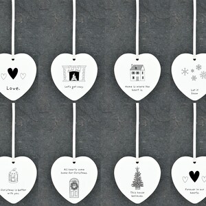 May include: Set of eight white ceramic heart-shaped ornaments with black and white illustrations and text. The ornaments feature phrases like "Love", "Let's get cozy", "Home is where the heart is", "Let it snow", "Christmas is better with you", "All hearts come home for Christmas", "This house believes", and "Forever in our hearts".
