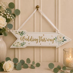 May include: A white arrow-shaped wooden sign with the gold text Wedding this way, decorated with painted white roses and green leaves. The sign hangs from a white ribbon and features small gold hearts scattered across the surface.