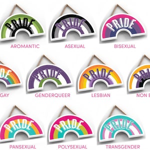 May include: Twelve rainbow-colored wooden signs with the word "PRIDE" in white lettering. Each sign features a different pride flag design, including aromantic, asexual, bisexual, gay, genderqueer, lesbian, non-binary, pansexual, polysexual, and transgender.