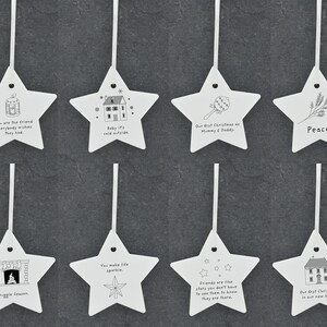 May include: Ten white star-shaped ornaments with black ink illustrations and text. The ornaments feature various holiday-themed designs, including a house, a fireplace, a star, a baby rattle, and a friend.