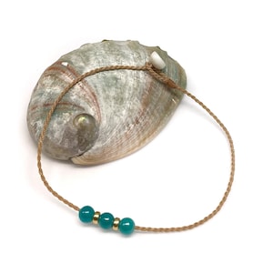 May include: A bracelet featuring a thin, braided tan cord with three smooth, round teal beads separated by two small, polished gold-tone metal spacers. The cord includes a small white bead near the adjustable knot closure mechanism.