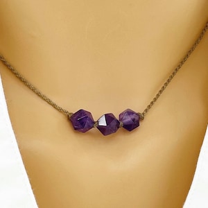 May include: A necklace featuring three faceted, deep purple amethyst beads strung on a thin, braided brown cord. The beads are arranged horizontally in the center of the cord, showcasing a geometric, multi-sided cut that reflects light.