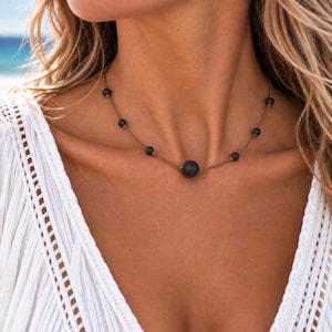 Peut inclure: Un collier composé d'un cordon fin marron clair sur lequel sont enfilées plusieurs petites perles sphériques sombres et poreuses, ainsi qu'une perle centrale plus grande, également sombre et poreuse, réparties le long du cordon.