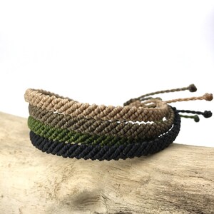 May include: Four macrame bracelets in shades of tan, olive green, and black are stacked on a piece of driftwood. The bracelets have a woven texture and adjustable ties. The neutral colors make them suitable for various outfits.