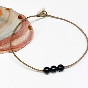 May include: A minimalist anklet featuring three glossy black beads strung on a thin, tan cord. The cord is knotted at the ends for adjustability. The anklet is displayed on a white surface with a seashell in the background.
