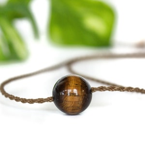 May include: A single spherical tiger's eye gemstone bead with a polished, glossy finish featuring brown and golden bands, threaded onto a thin, twisted brown cord that extends to both sides of the bead in a simple, minimalist necklace design.