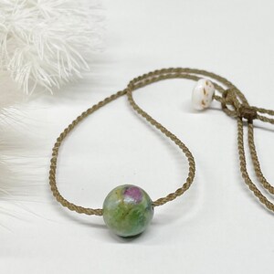 May include: A necklace featuring a single round, polished gemstone bead with mottled green and pink coloration, strung on a thin, braided tan cord. The cord includes a small, white, speckled cylindrical bead as a sliding adjustment mechanism.