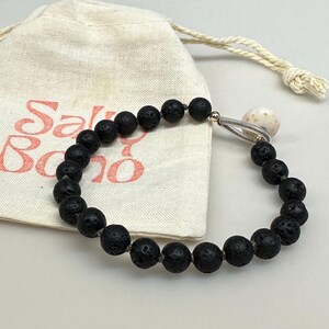 Pode incluir: Uma pulseira de contas de lava preta com um pingente prateado e uma pequena concha esbranquiçada. A pulseira está ao lado de uma bolsa com cordão com o texto "Salt Boho" em escrita coral.