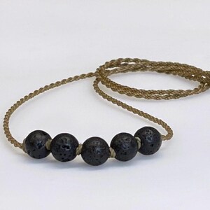 Peut inclure: Un collier composé de cinq perles rondes et poreuses en pierre de lave noire, enfilées sur un cordon torsadé de couleur beige. Les perles sont disposées en rangée horizontale au centre du cordon, qui forme une boucle pour le collier.