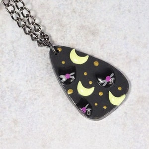 May include: A black teardrop-shaped pendant necklace with a black chain. The pendant features a repeating pattern of yellow crescent moons, gold dots, and black cat faces with pink noses.