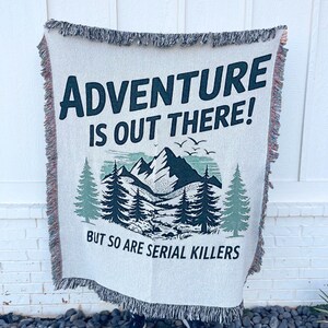 May include: Cream-colored woven throw blanket with the text "ADVENTURE IS OUT THERE! BUT SO ARE SERIAL KILLERS" in dark green. The design includes a mountain range, pine trees, and birds. The blanket has a fringed edge.