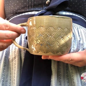Peut inclure: Une tasse en céramique faite à la main avec un glaçage beige et un motif de cercles répétés. La tasse a une anse incurvée et est tenue à deux mains. La tasse mesure environ 10 cm de haut.