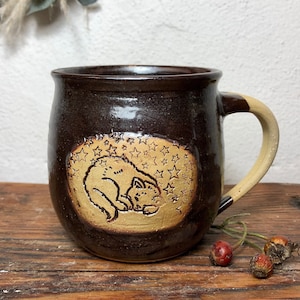 May include: A brown ceramic mug with a beige design of a cat sleeping under a sky full of stars. The mug has a rounded body and a handle.