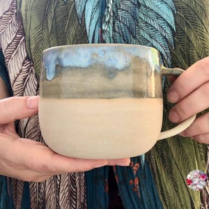 Può includere: Una tazza in ceramica fatta a mano con una forma arrotondata e un manico grande. La tazza presenta un design bicolore, con una base color crema e una sezione superiore che passa da un blu chiaro a un grigio-verde più scuro. La tazza è tenuta da una persona.