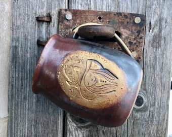 May include: A brown ceramic mug with a gold design of a bird with stars around it. The mug is hanging on a wooden door.