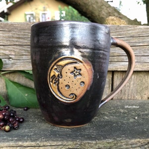 May include: A black ceramic mug with a brown crescent moon and star design. The mug has a handle and is sitting on a wooden surface.