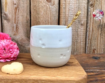 May include: A white ceramic mug with a floral design, a heart-shaped cookie, and a gold spoon on a wooden cutting board.