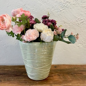 May include: A bouquet of pink and white flowers in a light green ceramic vase with a textured design. The vase is sitting on a wooden surface.