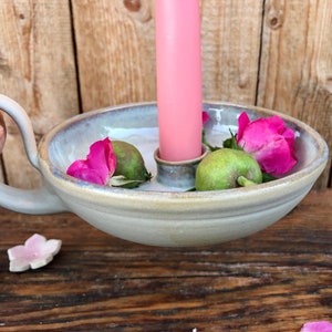 Könnte beinhalten: Ein Kerzenhalter aus Keramik mit Henkel, mit einer rosa Kerze, grünen Früchten und rosa Blütenblättern. Der Halter hat eine hellgraue Glasur.