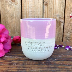 Könnte beinhalten: Eine zweifarbige Keramiktasse, oben rosa und unten cremefarben, mit den Worten "COFFEE THERAPY" in den cremefarbenen Bereich geätzt. Die Tasse steht auf einer Holzoberfläche, mit rosa Rosenblättern und einer Rose im Hintergrund.