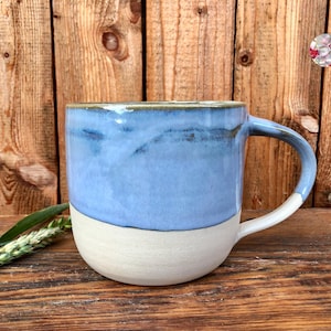 May include: A ceramic mug with a light blue glaze on the top half and a white glaze on the bottom half. The mug has a rounded shape and a handle.