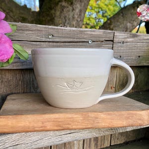 May include: A large, handmade ceramic mug with a boat design. The mug is two-toned, with a white upper half and a beige lower half. The boat design is etched into the beige section. The mug has a large handle and sits on a wooden board.
