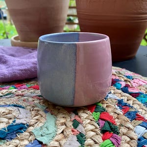 Può includere: Una piccola tazza in ceramica fatta a mano con un design bicolore. Una metà è blu-grigio tenue, l'altra rosa pallido. La tazza è appoggiata su un colorato tappetino intrecciato. Due vasi di terracotta sono sullo sfondo.
