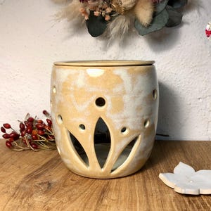 May include: A ceramic wax warmer with a white and beige glaze. The warmer has a floral design and a removable top. The warmer is sitting on a wooden surface.