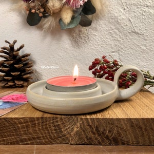 Könnte beinhalten: Ein weißer Kerzenhalter aus Keramik mit einem Griff. Der Halter hat eine kleine, runde Vertiefung für ein Teelicht. Ein rosa Teelicht brennt im Halter.