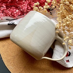 May include: A cream-colored ceramic mug with a rounded square shape and a curved handle. The mug is set on a white patterned cloth with dried flowers in the background. The mug has a glossy finish.