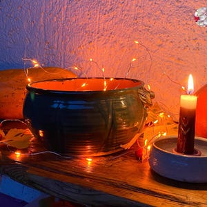 May include: A rustic, dark green ceramic bowl with an orange interior sits on a wooden surface, next to a lit candle in a white holder. A small pumpkin and string lights add to the autumn theme.