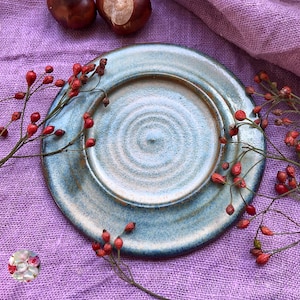 Può includere: Un piatto in ceramica fatto a mano con una glassa blu e marrone. Il piatto ha un design circolare con un centro rialzato. Bacche rosse e due castagne marroni sono disposte attorno al piatto su un tessuto viola.