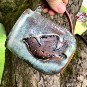 Pode incluir: Uma caneca de cerâmica artesanal com uma alça marrom e um relevo detalhado de um pássaro. A caneca apresenta uma mistura de esmalte azul, marrom e branco, criando uma aparência rústica e artística. O design do pássaro é uma característica proeminente.