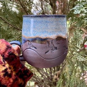 Könnte beinhalten: Eine handgefertigte Keramiktasse mit blau-brauner Glasur. Die Tasse hat ein geprägtes Design von Vögeln im Flug. Der Griff ist dunkelbraun. Die Tasse wird vor einem grünen Hintergrund gehalten.