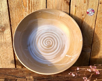 May include: A large, round, ceramic bowl with a white spiral design on a beige background. The bowl is sitting on a wooden surface.